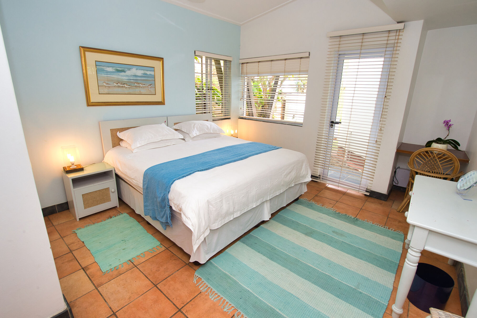 This is a room with one sky blue wall, and the remaining 3 white walls. There is one double bed, which consists of white linen and a blue throw-over. The roller blinds are white. There is a door to the right of the image, which has blinds in front of it. The door itself is transparent. There is a small turquoise mat next to the bed, and one large turquoise mat in front of the bed.