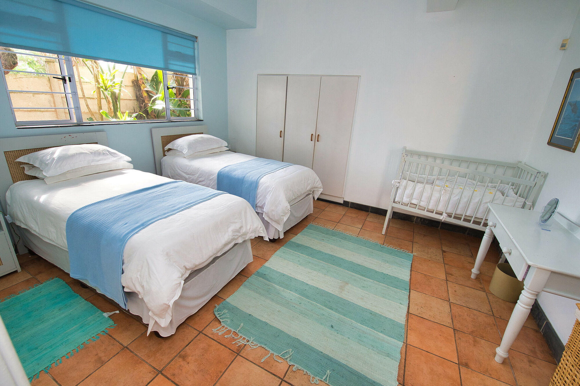This is a room with one sky blue wall, and the remaining 3 white walls. There are two single beds. Both beds consisting of white linen and a blue throw-over. There is a cot for a baby. The roller blinds are also blue.