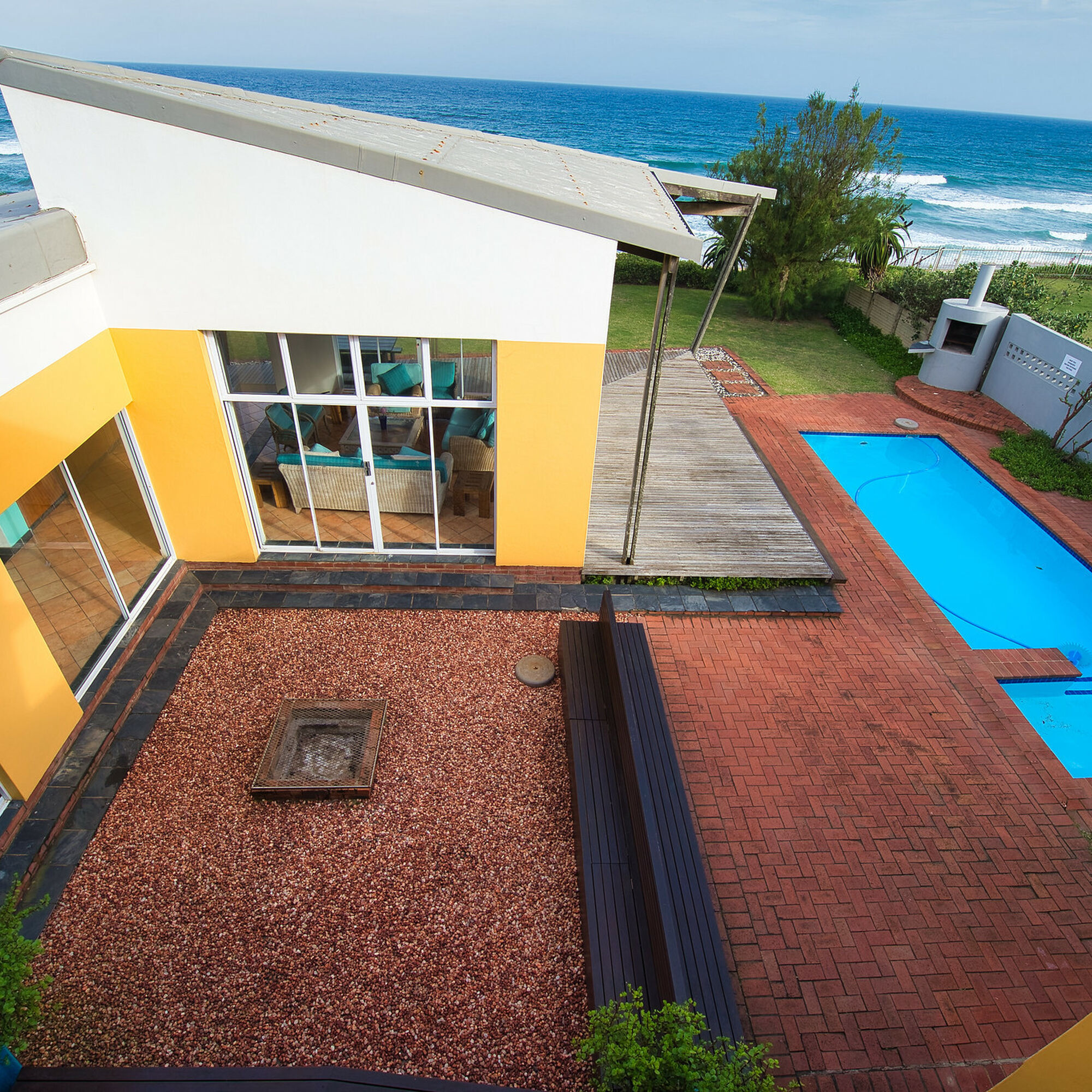 A bird's eye view of the holiday home. This part of the home has yellow walls . Trees surround the home. There are pebbles towards the braai area. The pool is on the right side, surrounded by laid bricks. The ocean is in the distance. The sky is a clear pale blue.