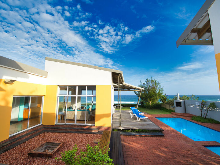 The braai area is on the left, the floor is made up of pebbles. The home is directly in front of the viewer. It has yellow and white walls. The pool is towards the right side of the image. The sky is blue, and is partially cloudy.