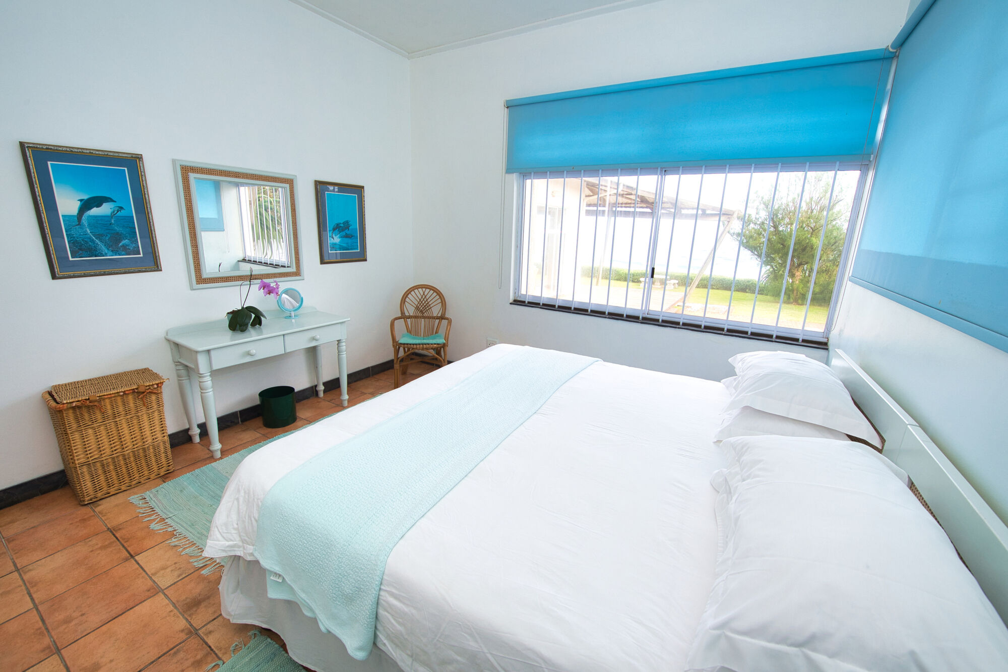 This is a room with a double sized bed. The bed has white linen and a sky blue throw-over. A dressing table and a mirror are found between two blue paintings of dolphins. The window roller blinds are also blue.